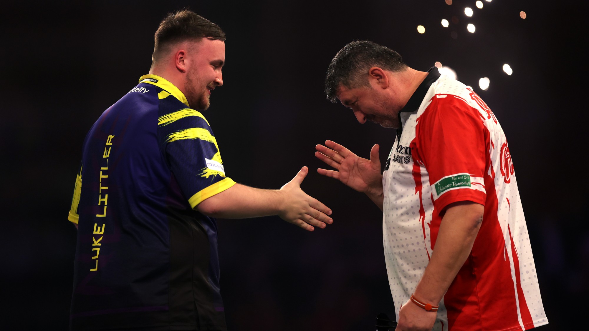 Luke Littler (balra) és Mensur Suljovic kezet ráznak, miután előbbi nyert a dartsvilágbajnokságon, Londonban.