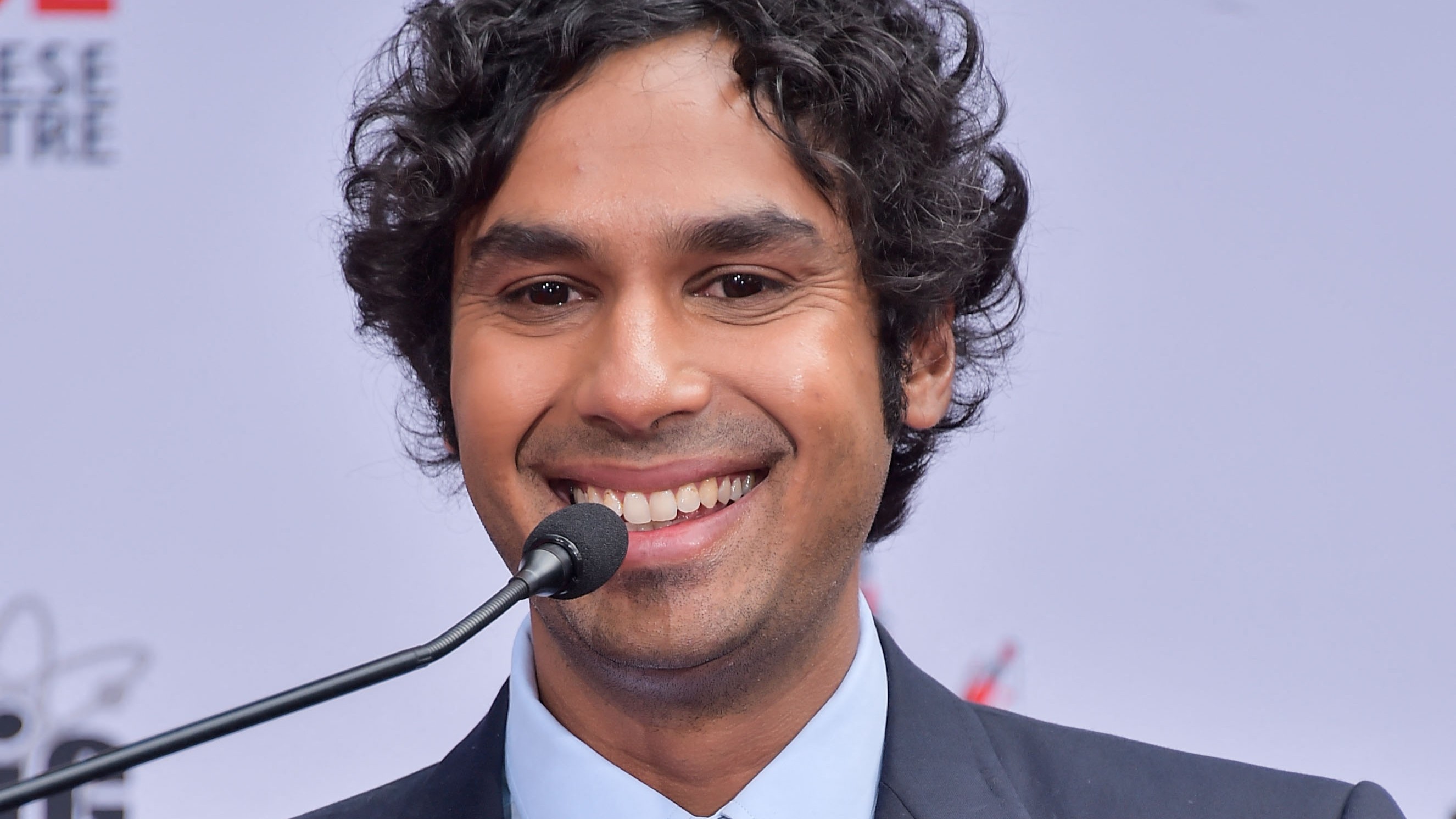 Kunal Nayyar egy mikrofon mögött állva mosolyog, egy világos háttér előtt