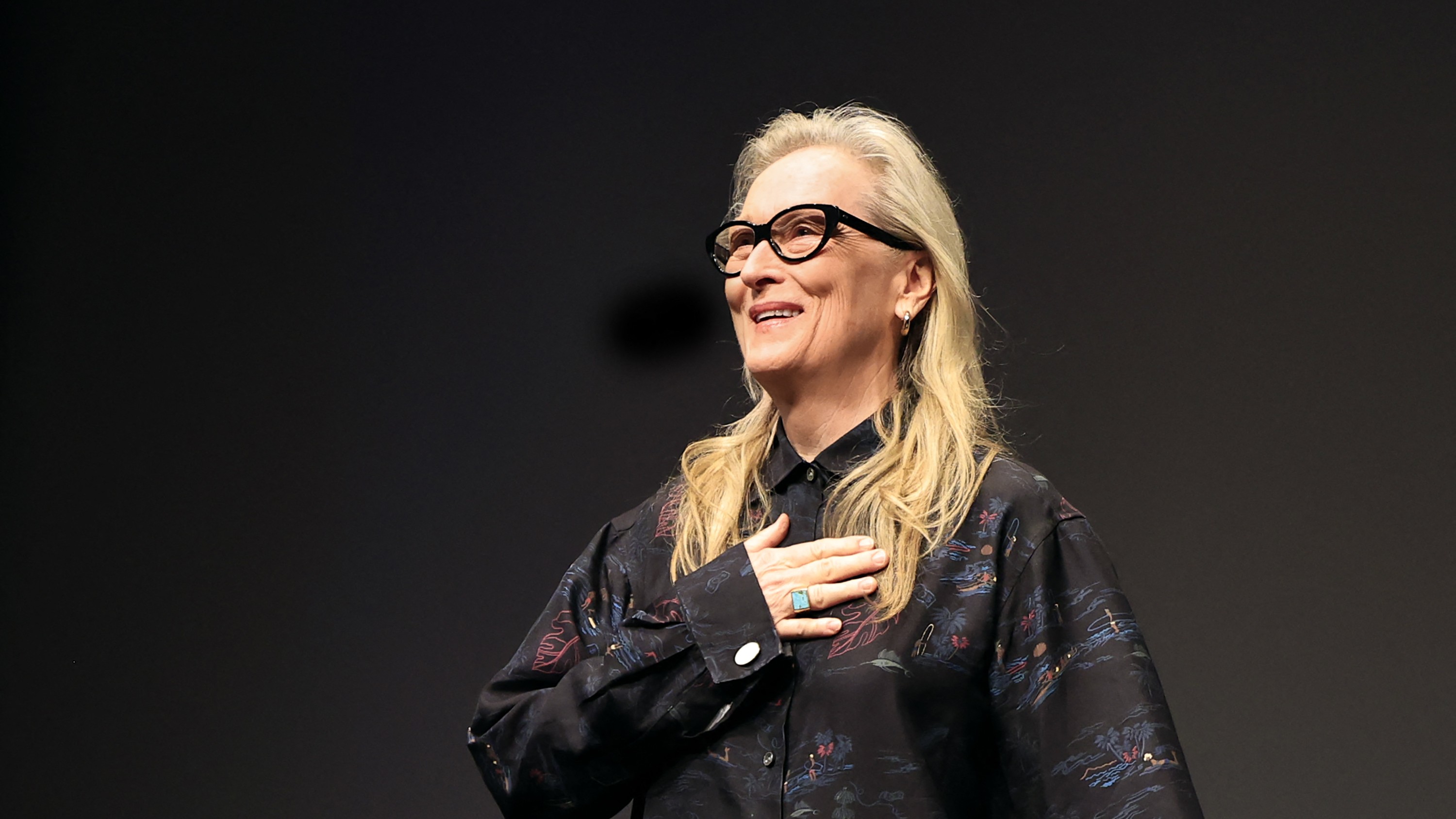 Meryl Streep mellkasra tett kézzel mosolyog egy sötét háttér előtt