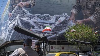 Két idős iráni férfi áll egymás mellett egy hatalmas óriásplakát alatt, amelyen egy mesterséges intelligencia által generált kép látható, 2026. április 8-án Teherán belvárosában, Iránban.