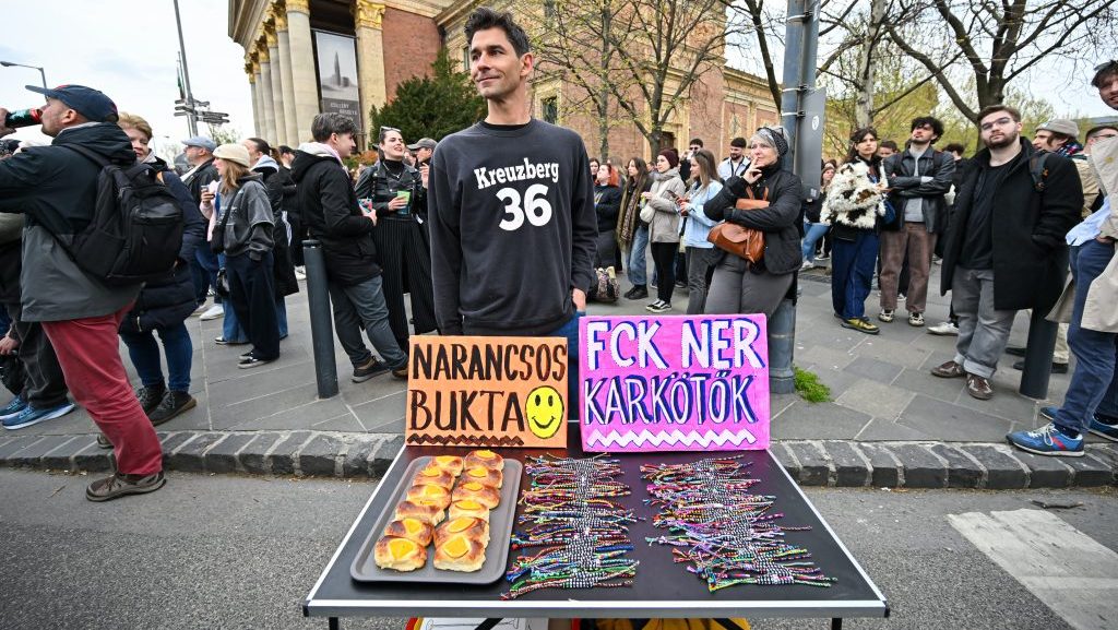 Egy férfi narancsos buktát és kormányellenes karkötőket árul a Rendszerbontó Nagykoncerten a budapesti Hősök terén, 2026. április 10-én.