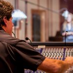 Male caucasian middle aged recording engineer in a recording studio sitting at a multi channel console in front of an isolated room with a pretty young woman singing into a microphone