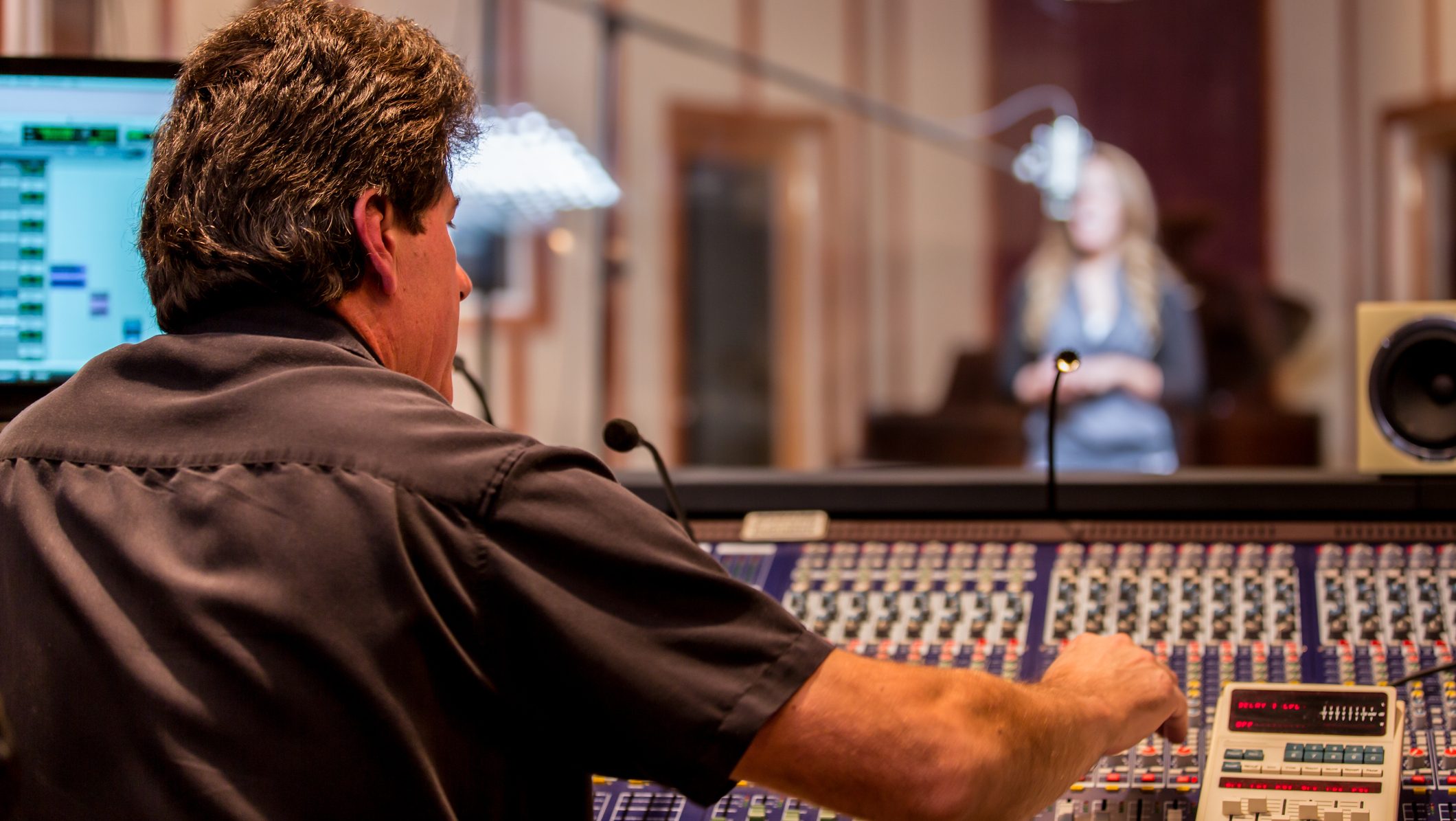 Male caucasian middle aged recording engineer in a recording studio sitting at a multi channel console in front of an isolated room with a pretty young woman singing into a microphone