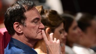 Close-up side profile of a man in a blue shirt with his finger on his lips, appearing thoughtful in a crowded audience.