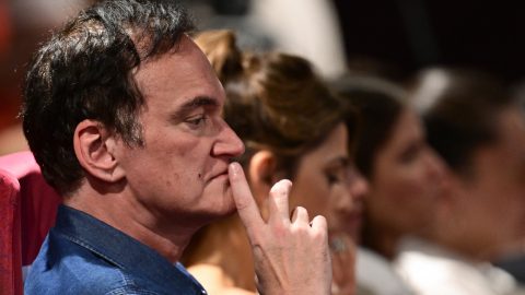 Close-up side profile of a man in a blue shirt with his finger on his lips, appearing thoughtful in a crowded audience.