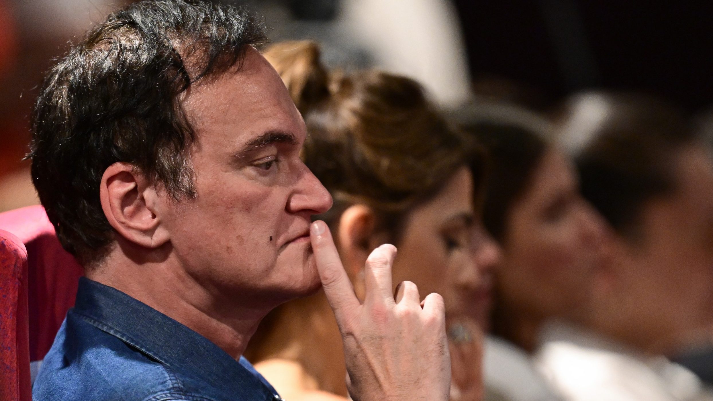 Close-up side profile of a man in a blue shirt with his finger on his lips, appearing thoughtful in a crowded audience.