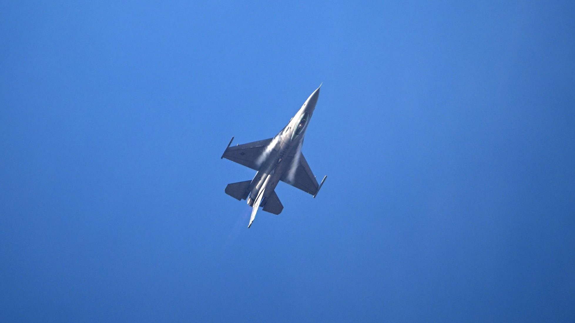 Fighter jet seen from below, gliding through clear blue sky with light vapor trails behind it.