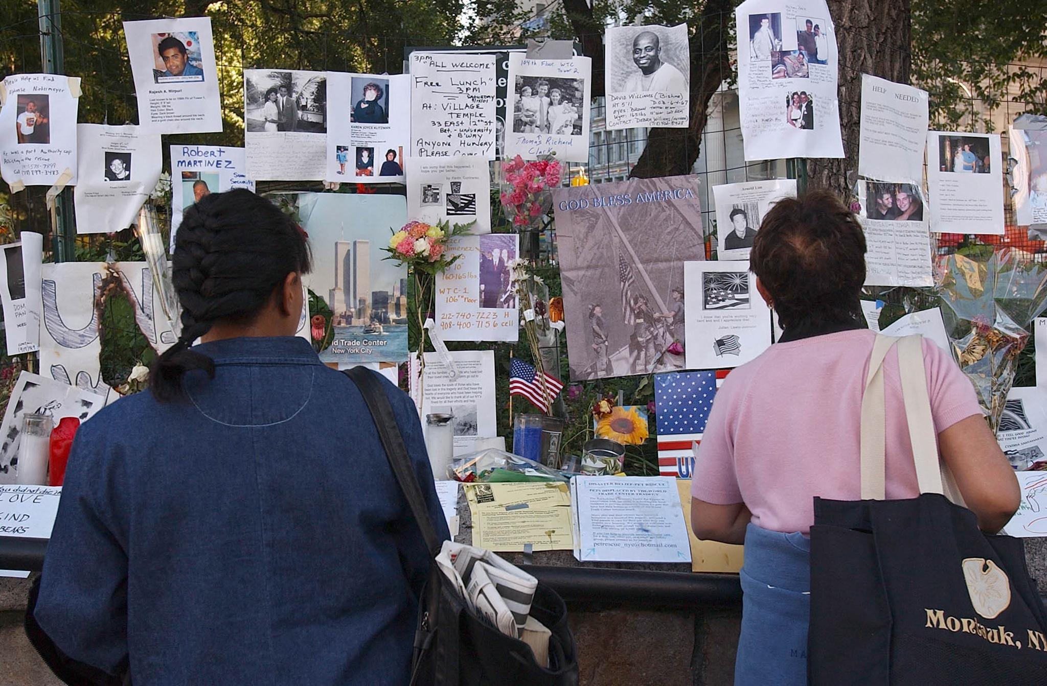 Two people with bags stand before a wall covered in photos, notes, and flowers at a memorial display.