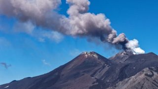 Volcano with dark rocky slopes emitting a tall plume of ash into a clear blue sky.
