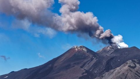 Volcano with dark rocky slopes emitting a tall plume of ash into a clear blue sky.