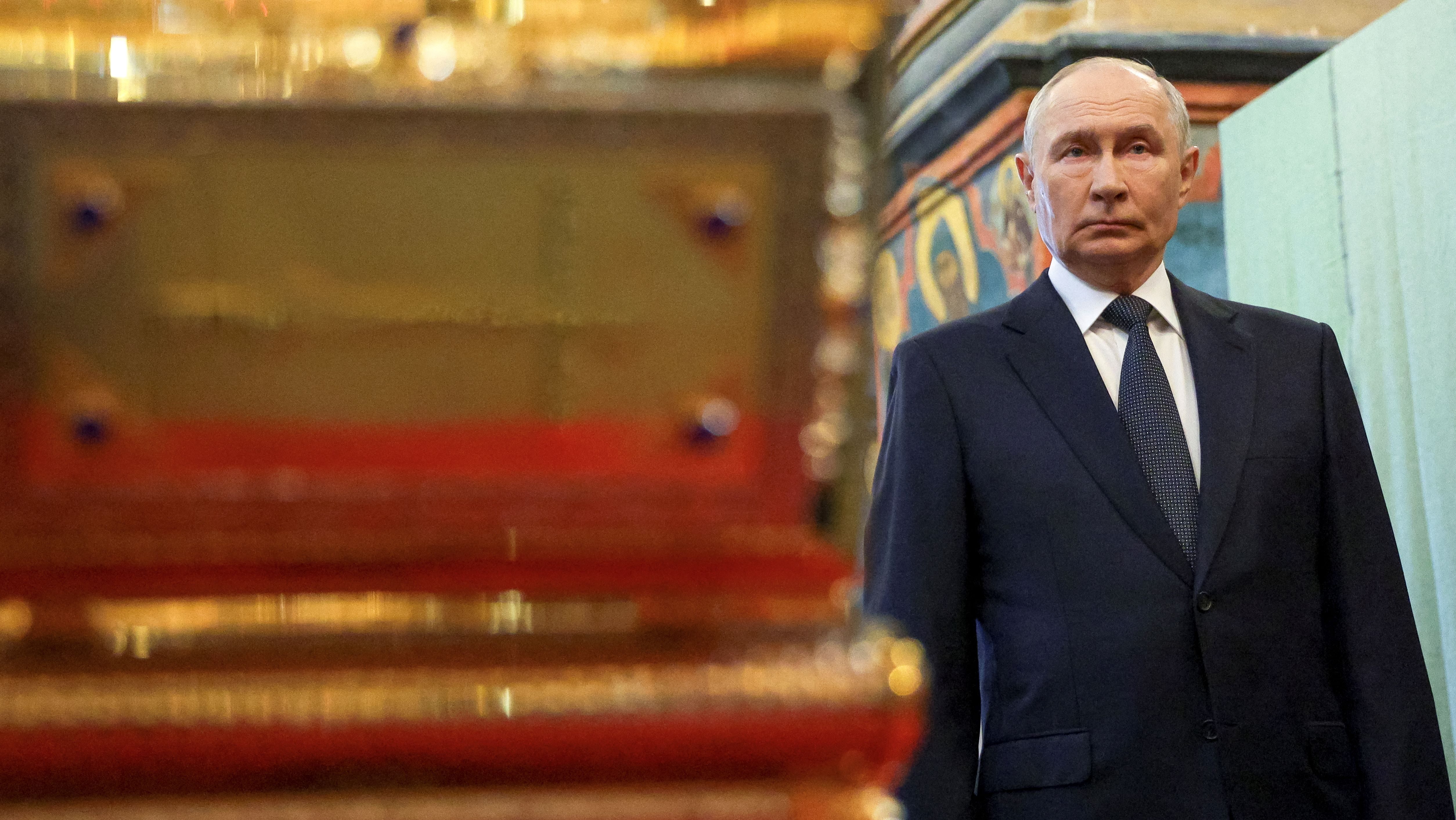 Portrait of Vladimir Putin in a dark suit and tie, standing beside a colorful mural in a formal setting.