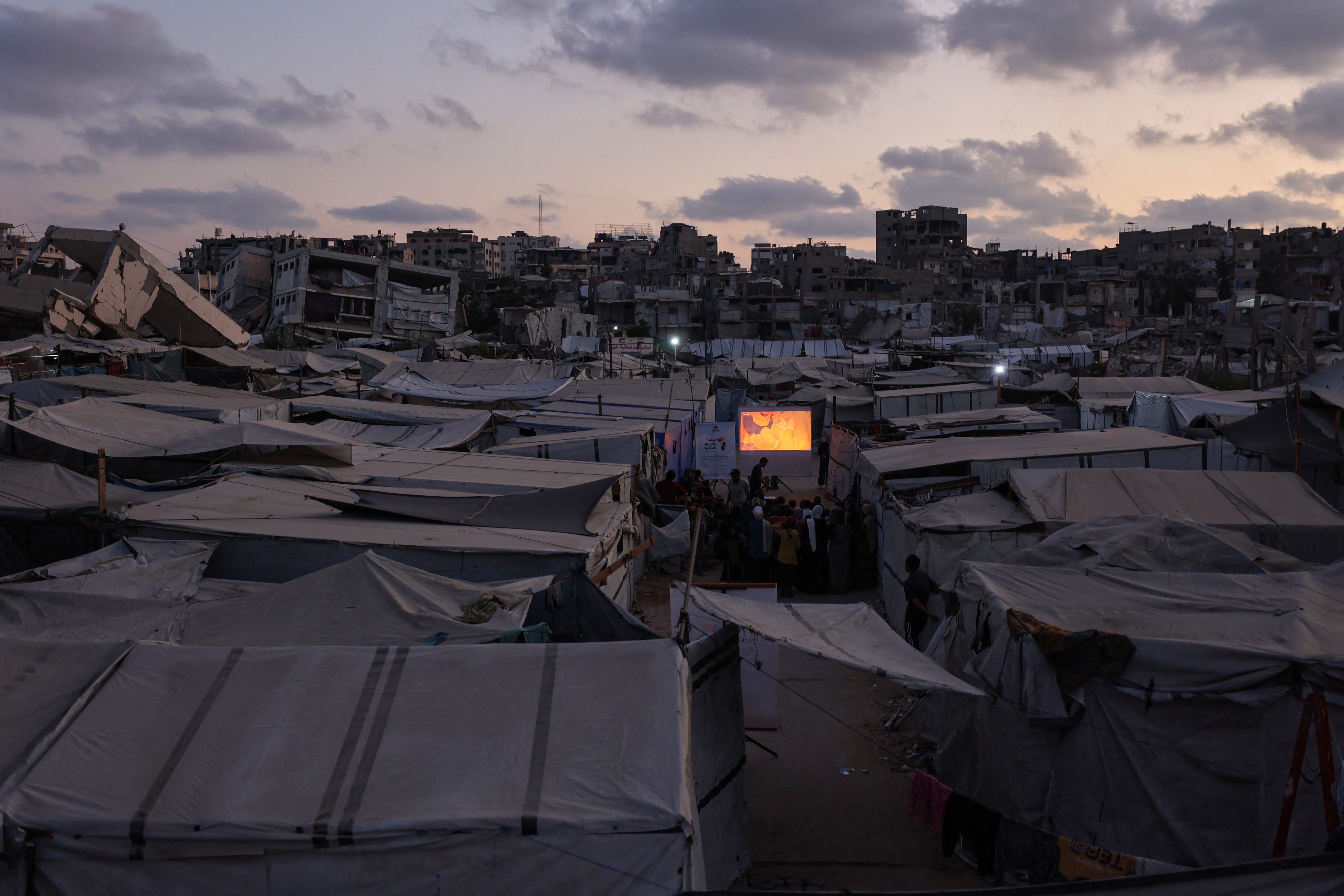 Dusk over a large refugee camp with rows of tents and damaged buildings in the background; a projector screen glows orange as people gather nearby.