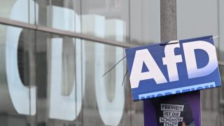 Blue and white political-style sign with large white letters clipped to a glass storefront, reflected city windows visible in the background, plus a purple poster beneath bearing German text about liberty.