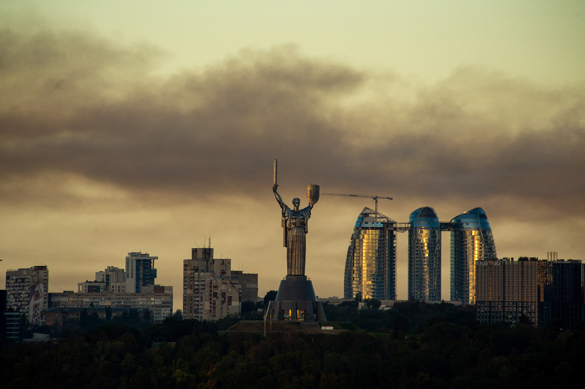 Statue of a woman with a raised sword overlooking a city skyline with futuristic glass buildings and a construction crane at sunset.
