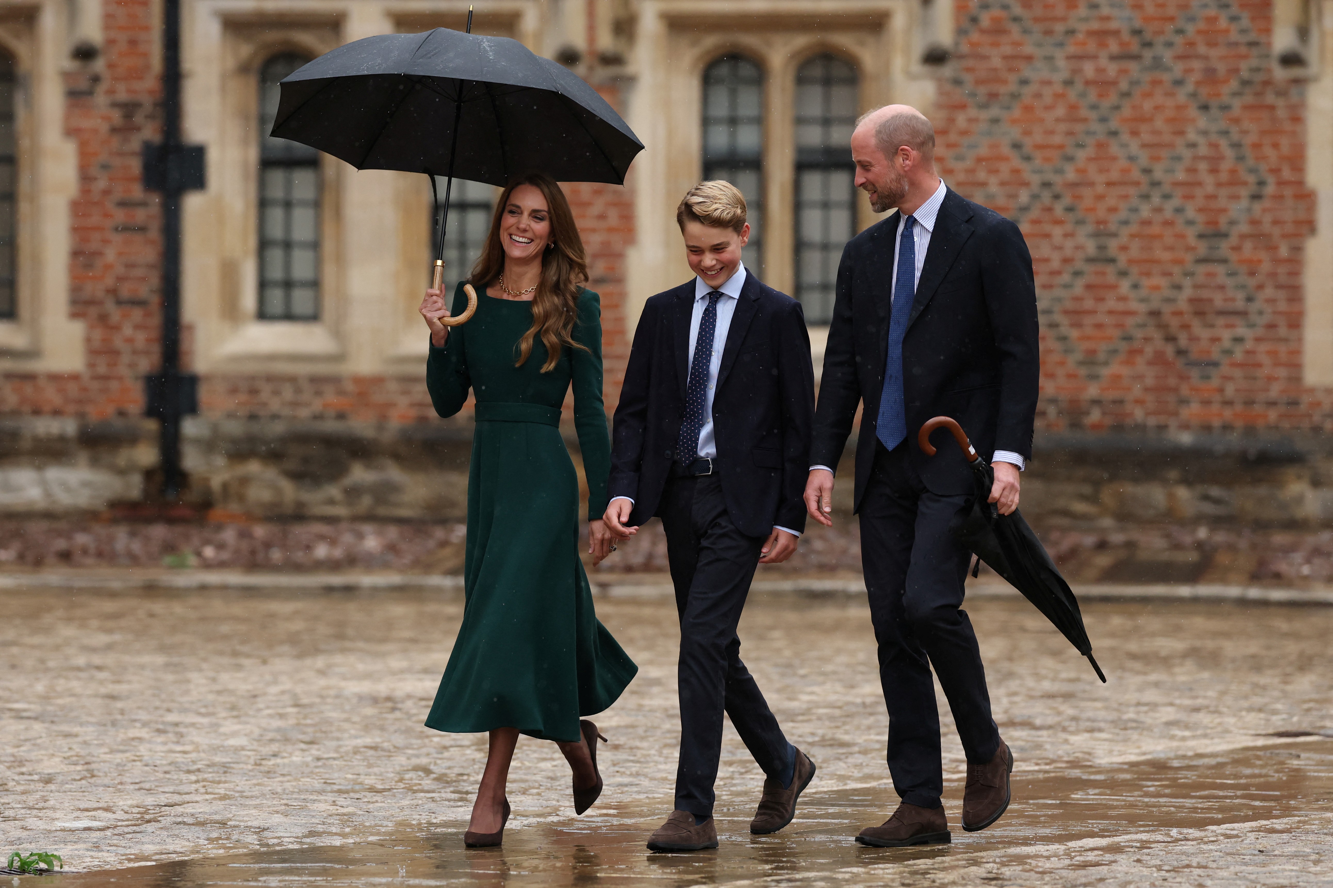 Three formally dressed people walking in the rain, smiling, one holding a large black umbrella.