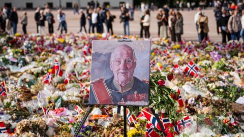 Portrait painting on an easel at a memorial, surrounded by flowers and Norwegian flags with a crowd in the background.