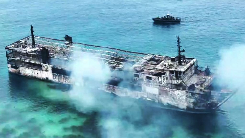 Wrecked ship floating in clear turquoise water with smoke rising from its deck, a small boat nearby in the distance.