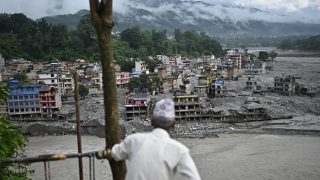 A person in a white shirt and patterned cap stands at a railing, looking over a mud-covered hillside town with ruined multi-story buildings and rubble, mountains in the cloudy background.
