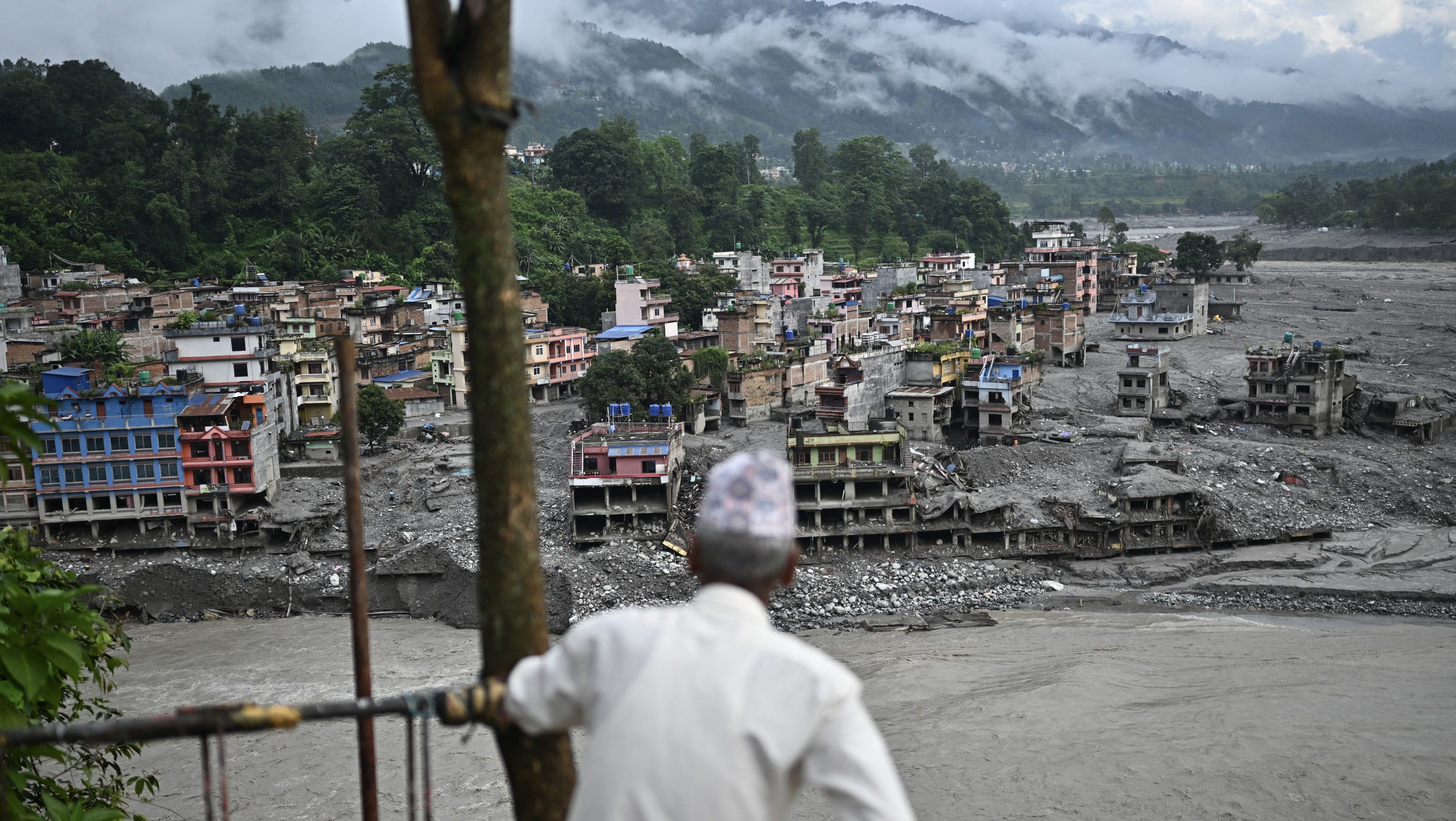 A person in a white shirt and patterned cap stands at a railing, looking over a mud-covered hillside town with ruined multi-story buildings and rubble, mountains in the cloudy background.