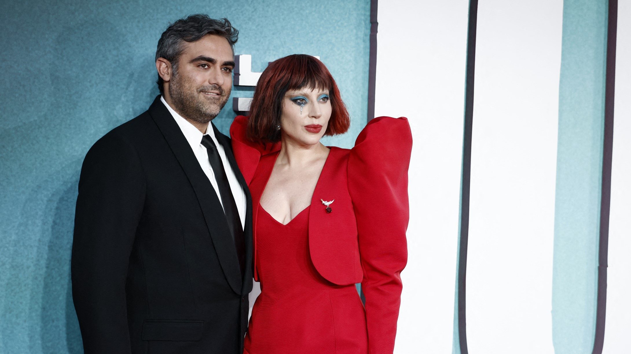 A man in a black suit and white shirt stands beside a woman in a red gown against a teal backdrop at a formal event on the red carpet, posing for photos.
