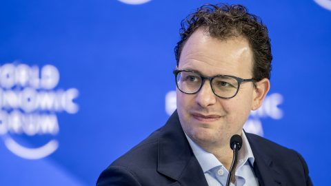 Dario Amodei wearing glasses and a dark suit speaks into a microphone at a press conference against a blue backdrop.