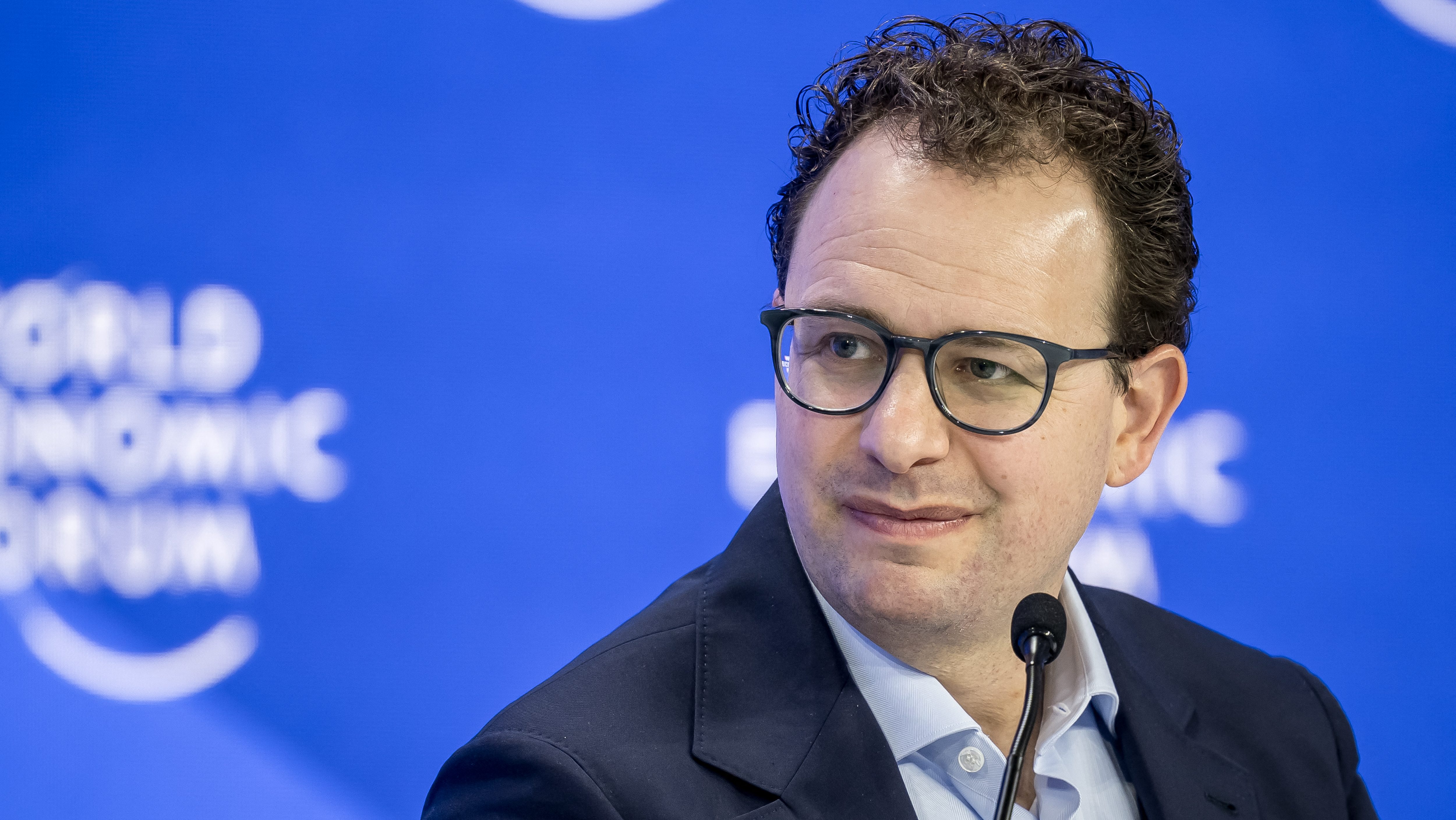 Dario Amodei wearing glasses and a dark suit speaks into a microphone at a press conference against a blue backdrop.