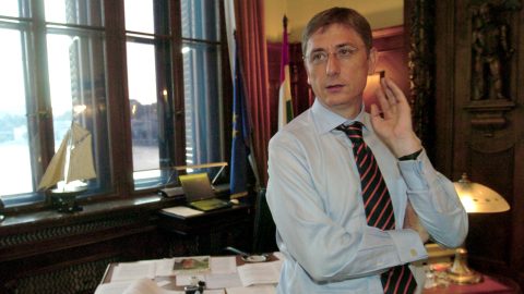 Man in a light blue shirt and striped tie cupping his ear, standing beside a cluttered desk in a wood-paneled office with windows and flags behind him.