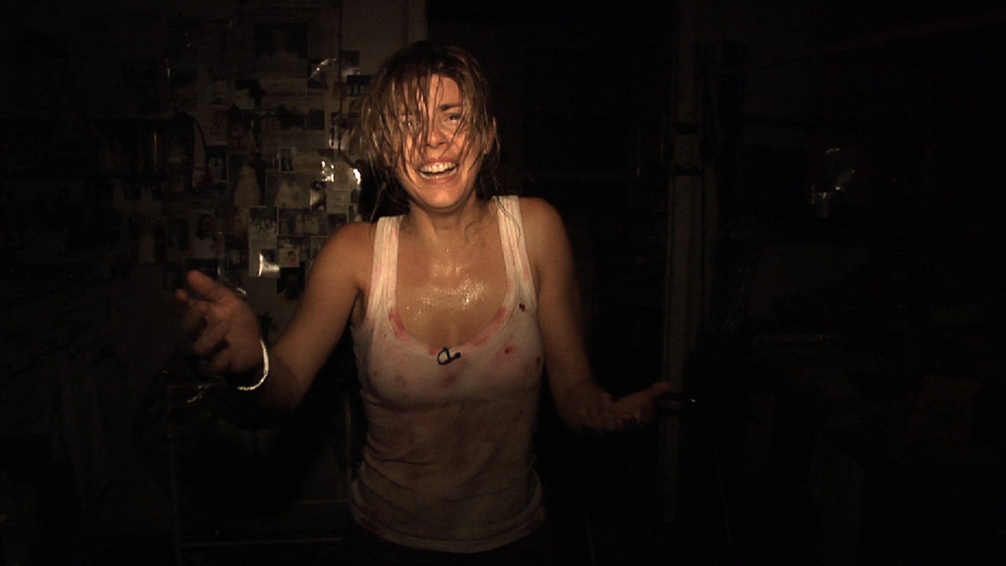 Woman with wet hair and a stained white tank top stands in a dark room, arms outstretched and smiling.