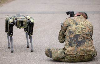 Quadrobot with camouflaged body standing on pavement beside a kneeling soldier operating a camera-equipped helmet unit.