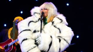 Performer in a white fur coat with black stripes singing into a microphone on stage, with a guitarist and blue stage lights in the background.