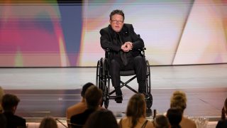 Man in a black suit seated in a wheelchair on a stage, addressing an audience with a colorful abstract backdrop behind him.