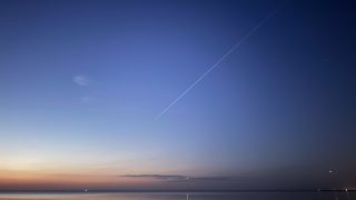 Twilight sky over a calm sea with an orange horizon, blue gradient above, and a diagonal airplane contrail crossing the scene.