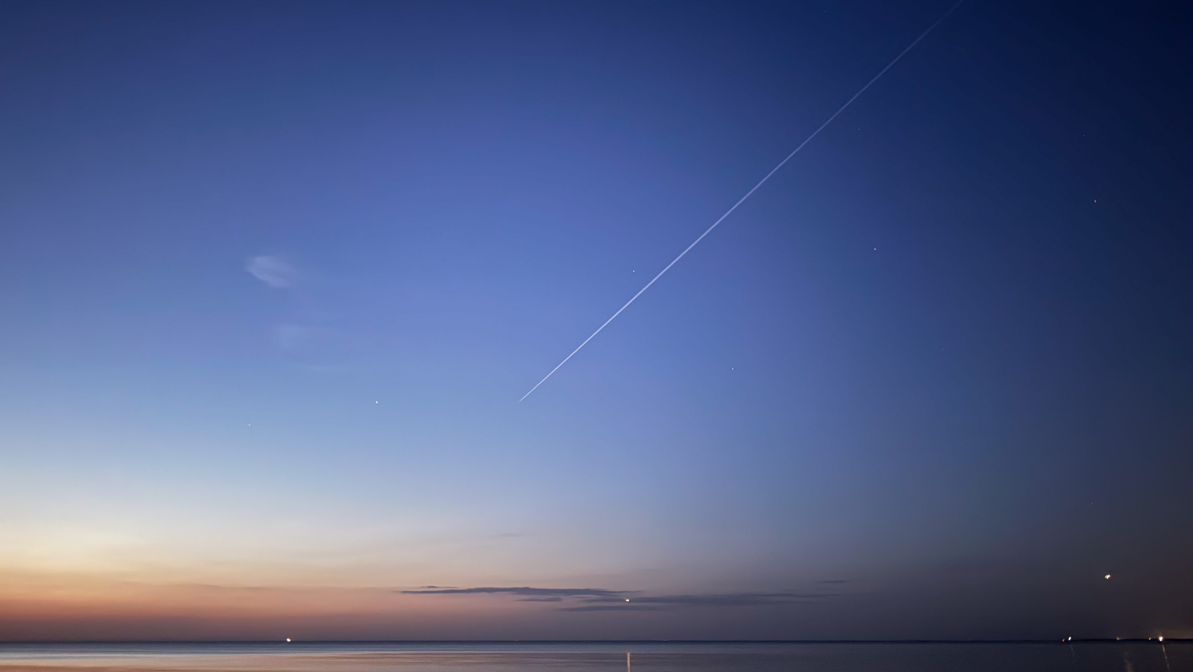Twilight sky over a calm sea with an orange horizon, blue gradient above, and a diagonal airplane contrail crossing the scene.