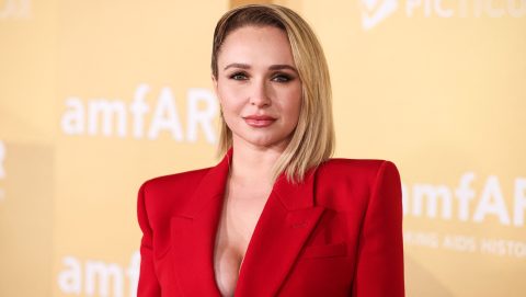 Portrait of a blonde woman in a red blazer at an amfAR event, looking at the camera against a pale yellow backdrop.