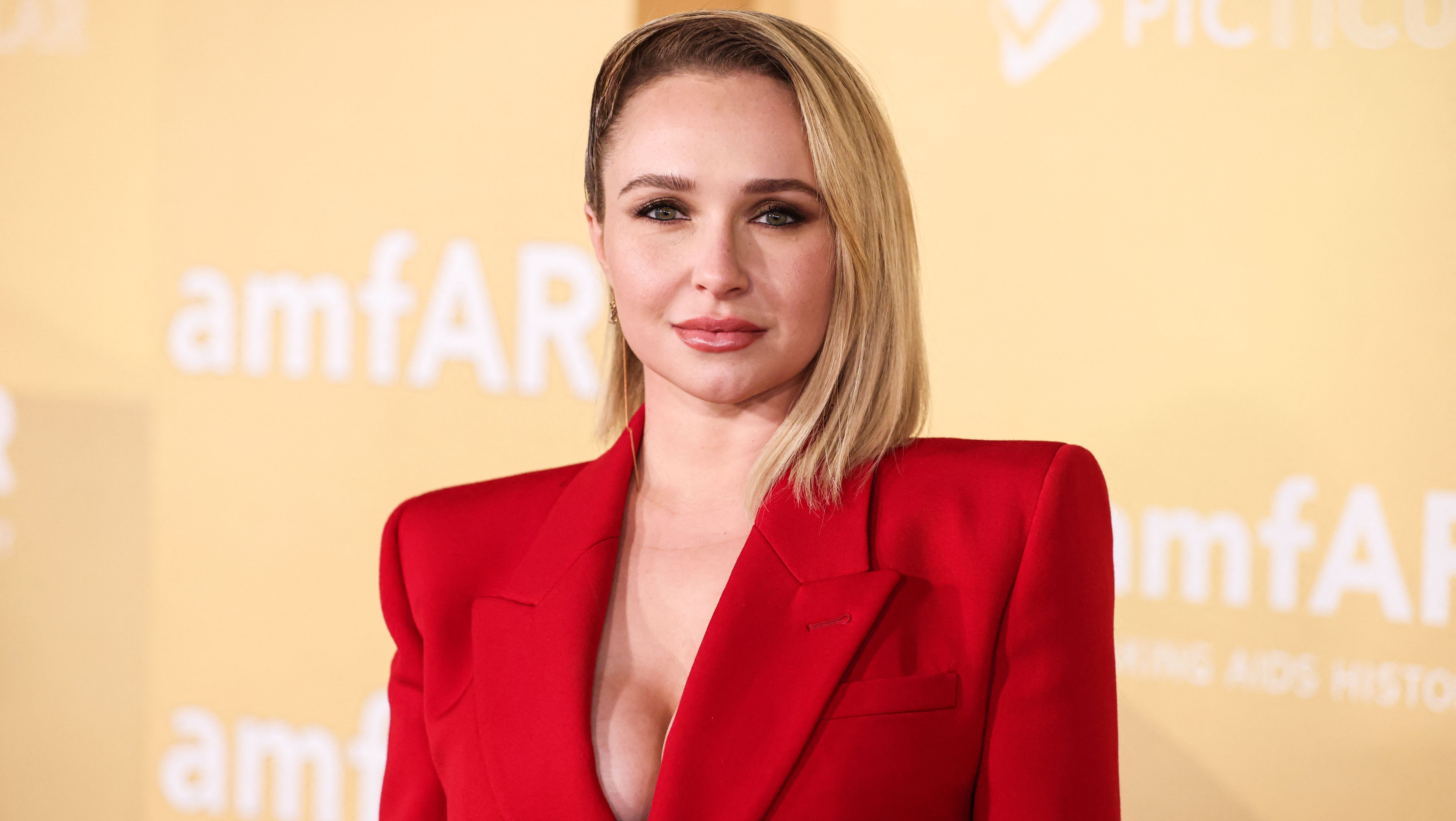 Portrait of a blonde woman in a red blazer at an amfAR event, looking at the camera against a pale yellow backdrop.