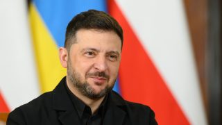 Close-up of a man with dark hair and a trimmed beard, wearing a black jacket, smiling with colorful blurred flag background behind him.