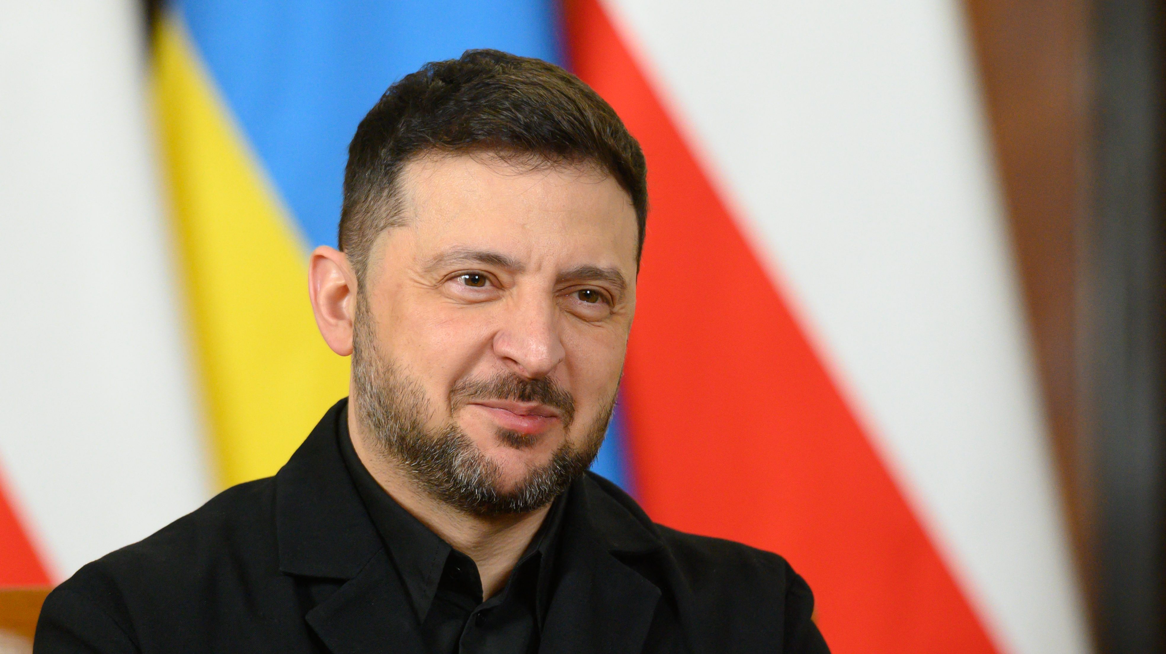 Close-up of a man with dark hair and a trimmed beard, wearing a black jacket, smiling with colorful blurred flag background behind him.