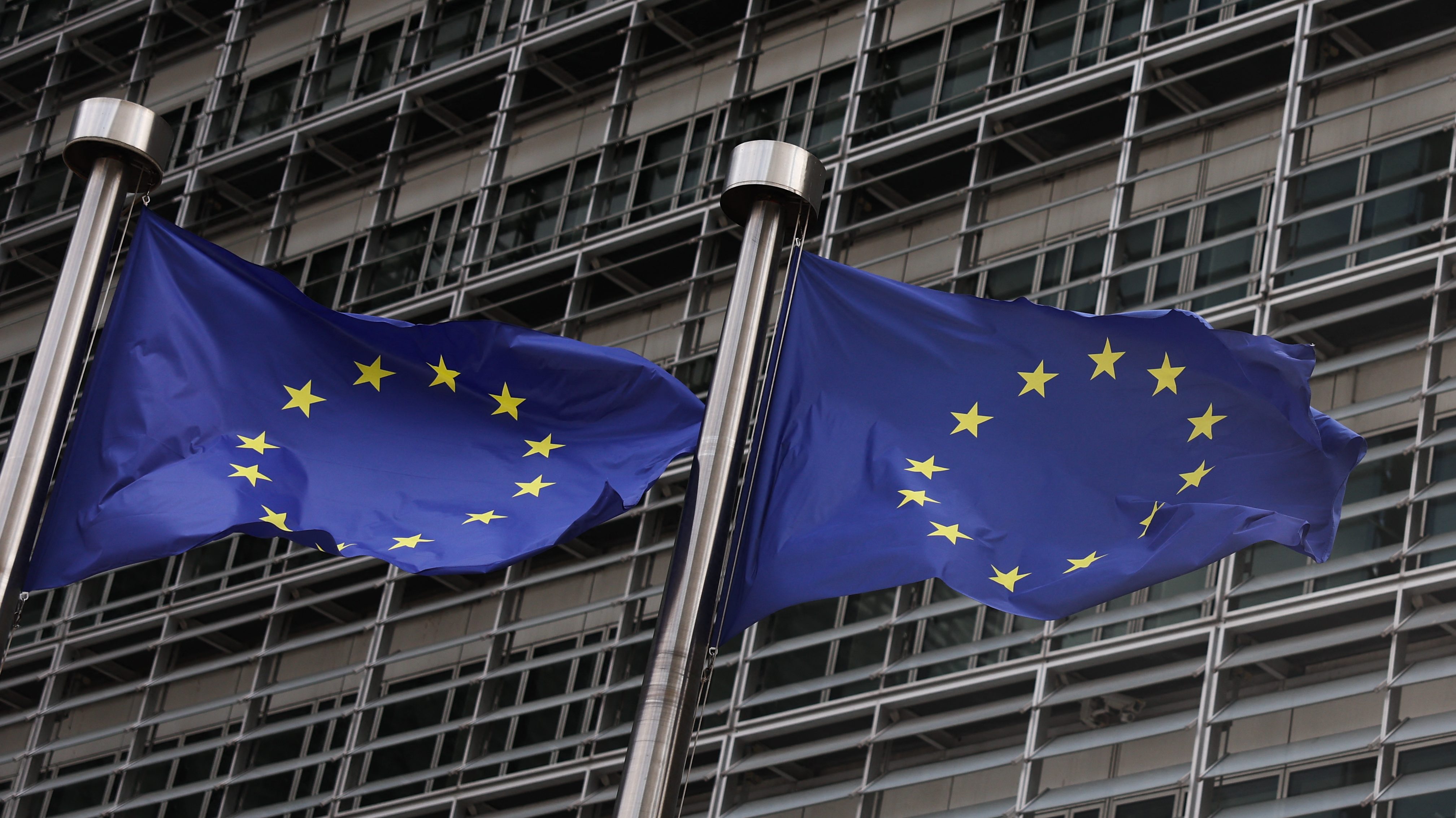 Two European Union flags on silver flagpoles waving in front of a modern glass office building rimmed with metal grids.
