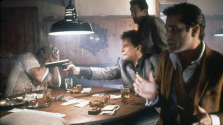 Tense scene in a bar: a man points a pistol at a seated man while two others stand nearby, watching intently around a cluttered table with money and drinks.