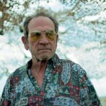 Older man wearing aviator sunglasses and a colorful Hawaiian-style shirt stands outdoors with trees in the background.