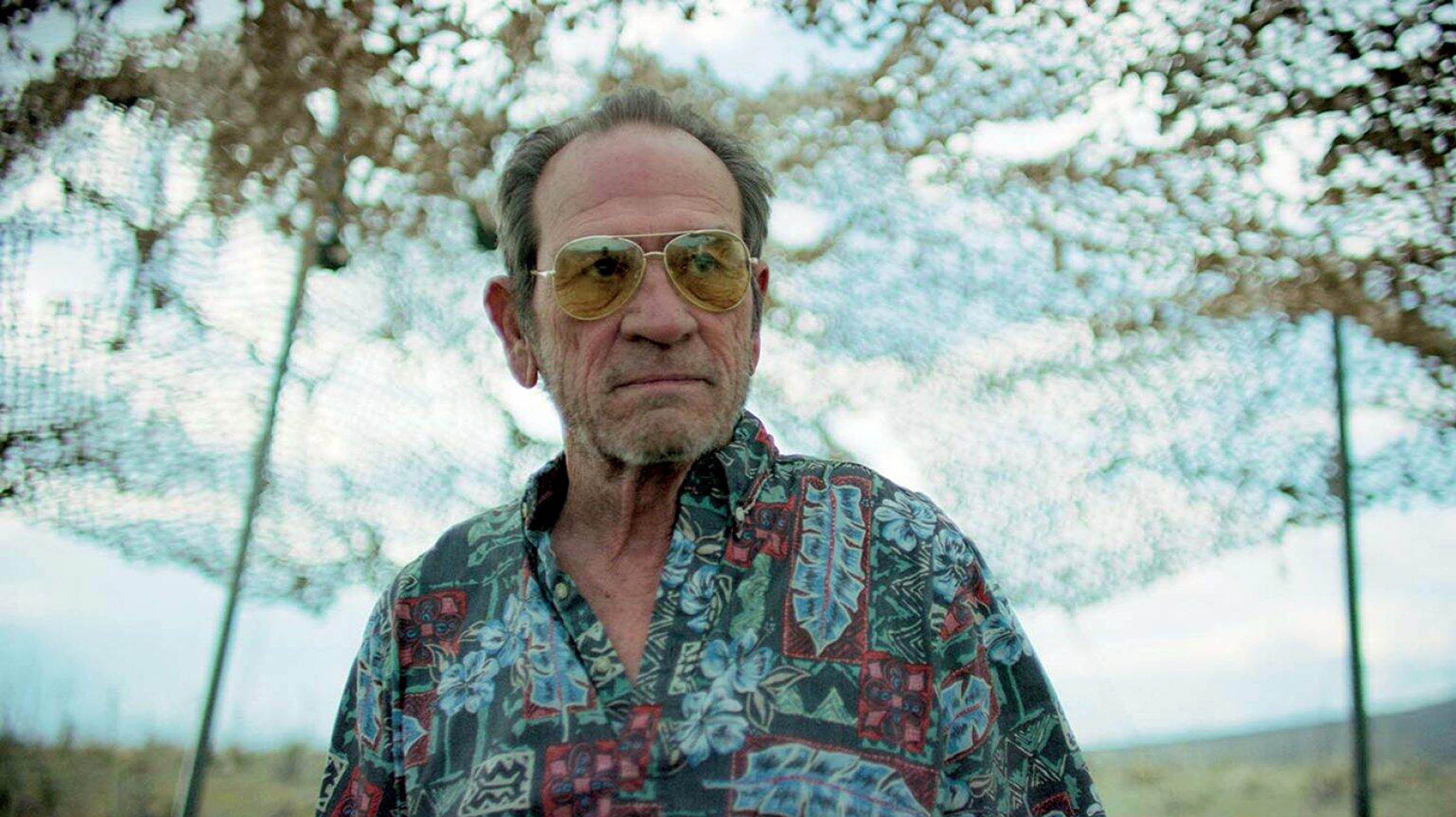 Older man wearing aviator sunglasses and a colorful Hawaiian-style shirt stands outdoors with trees in the background.