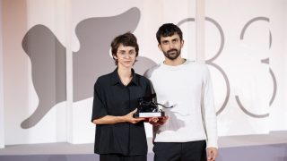 Two people pose with a trophy at an event; woman in black shirt and glasses stands beside a man in a light sweater against a patterned backdrop.