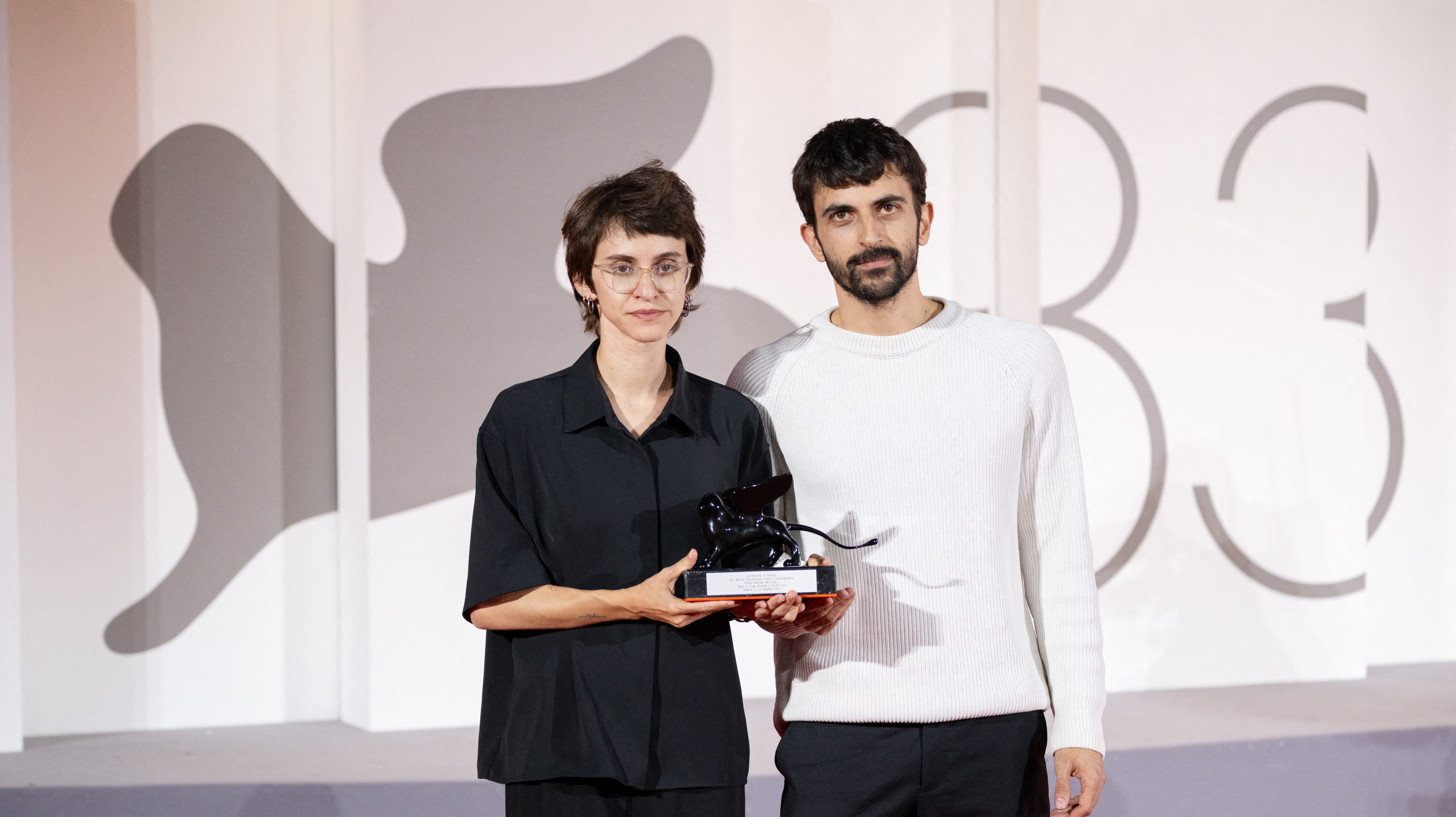 Two people pose with a trophy at an event; woman in black shirt and glasses stands beside a man in a light sweater against a patterned backdrop.