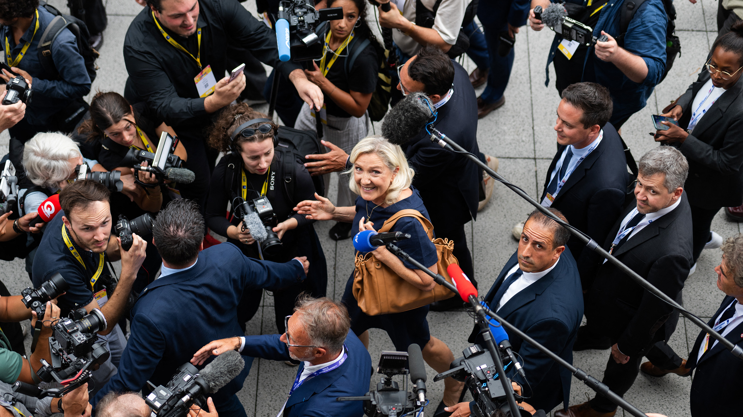 Overhead view of journalists with cameras surrounding a smiling blonde woman who speaks into multiple microphones.