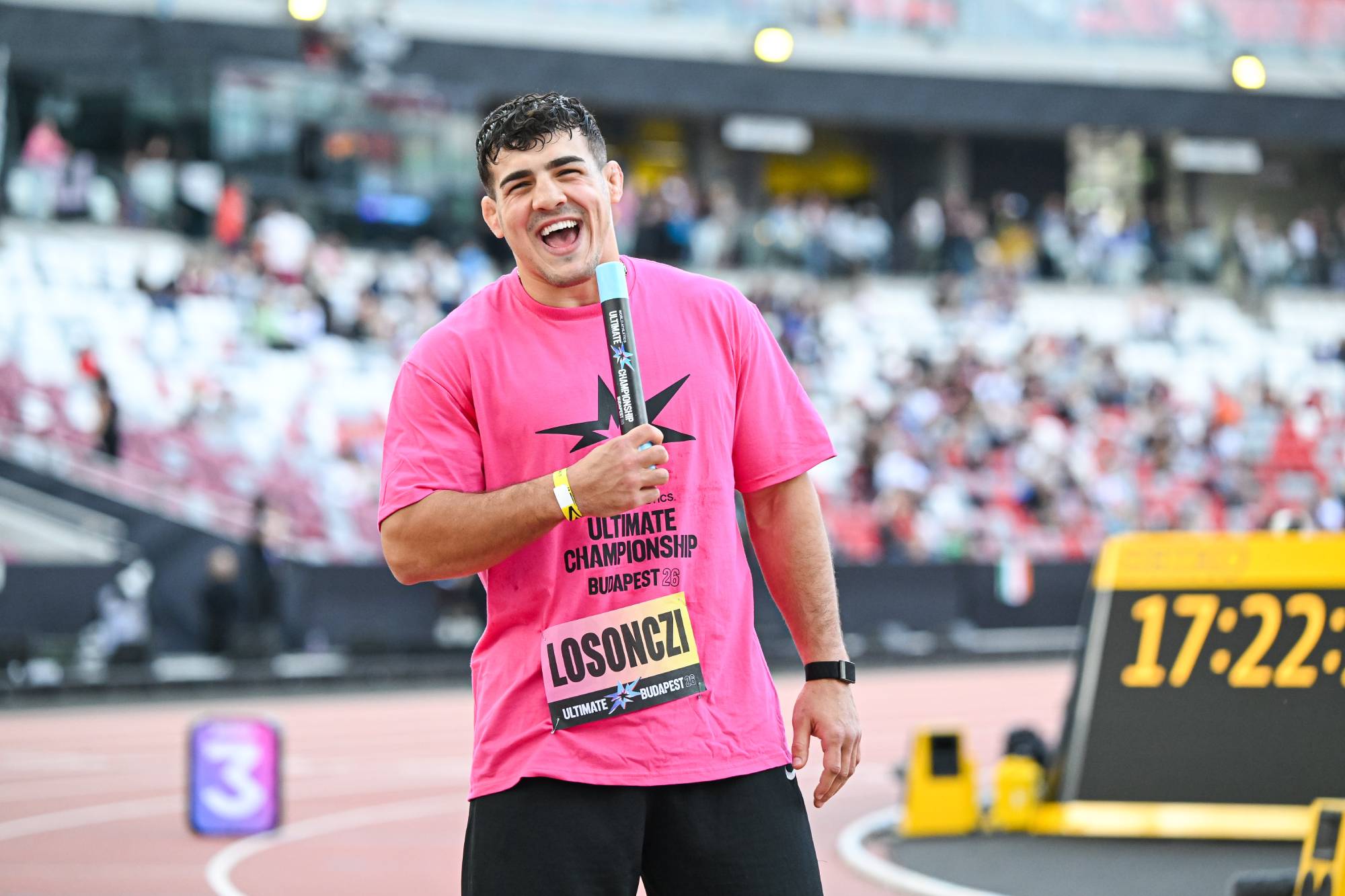 Male runner in a pink shirt smiles at the camera during a race at a stadium finish area, with a crowd and timing board in the background