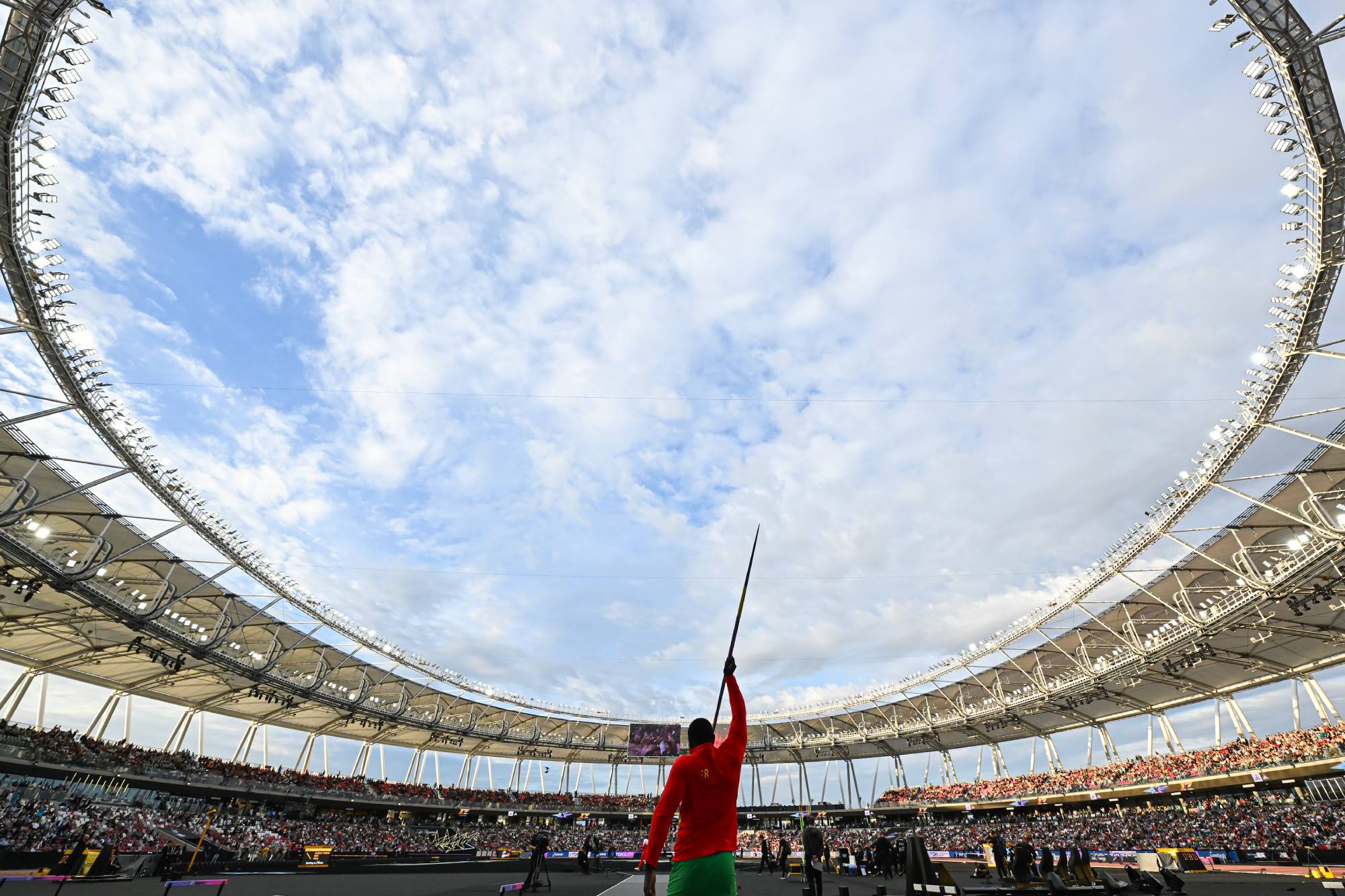 Athlete in a bright red outfit stands center stage inside a large circular stadium, holding a long red flag while a full audience watches from the stands.