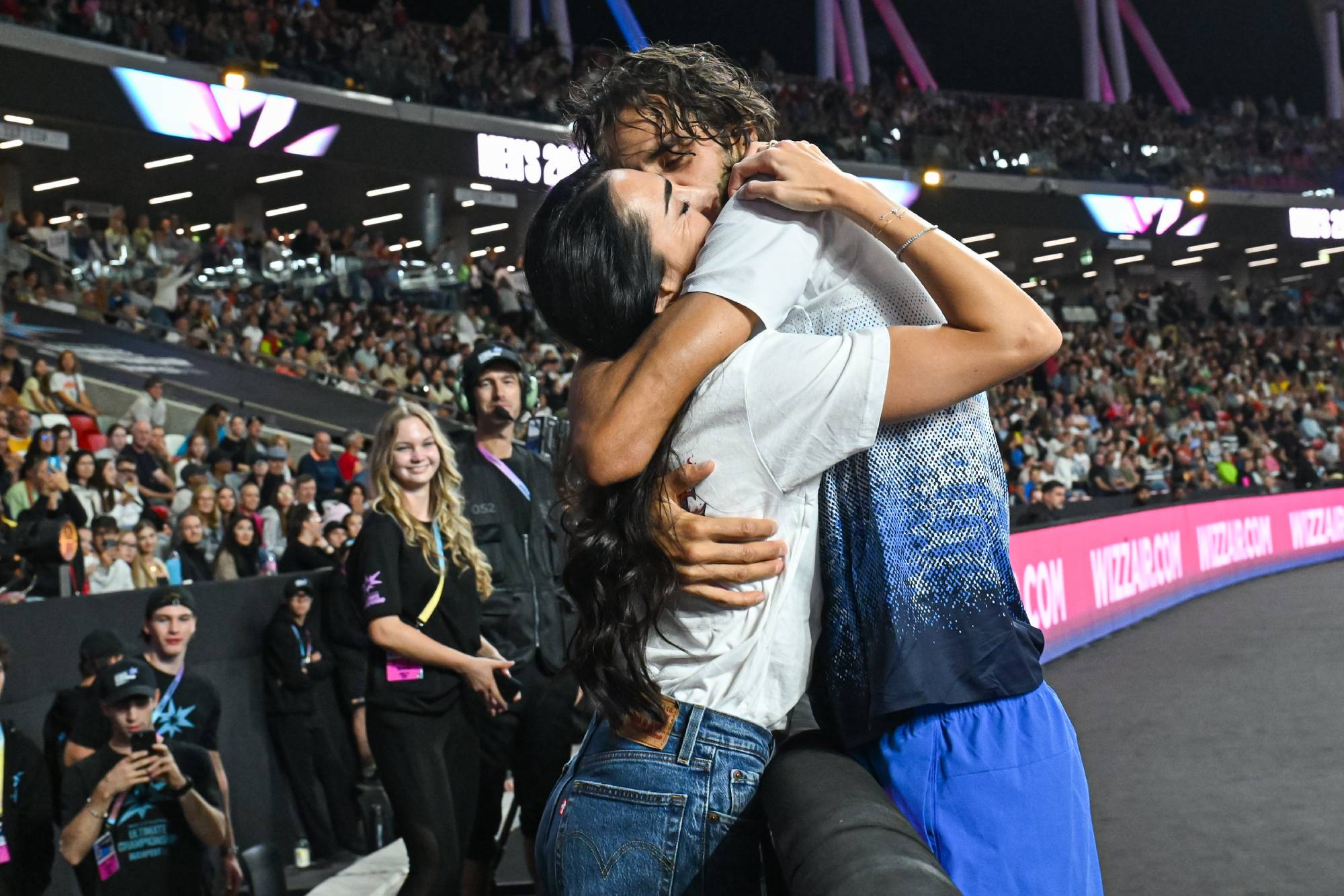 Two people hug tightly in a packed arena, with fans in the stands watching the moment behind them.
