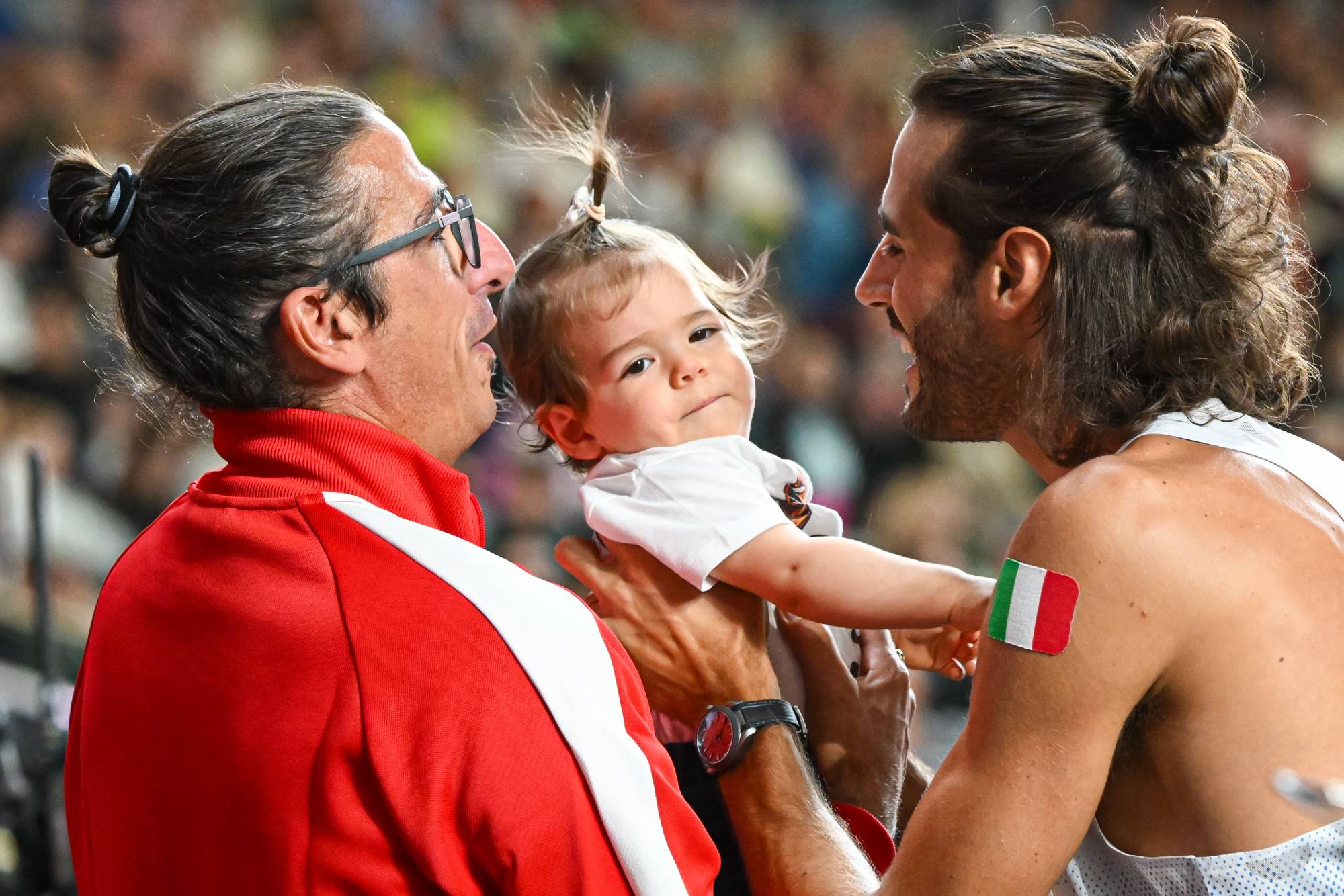 Man in a red jacket holds a baby while another man with long hair smiles at them in a crowded sport event.