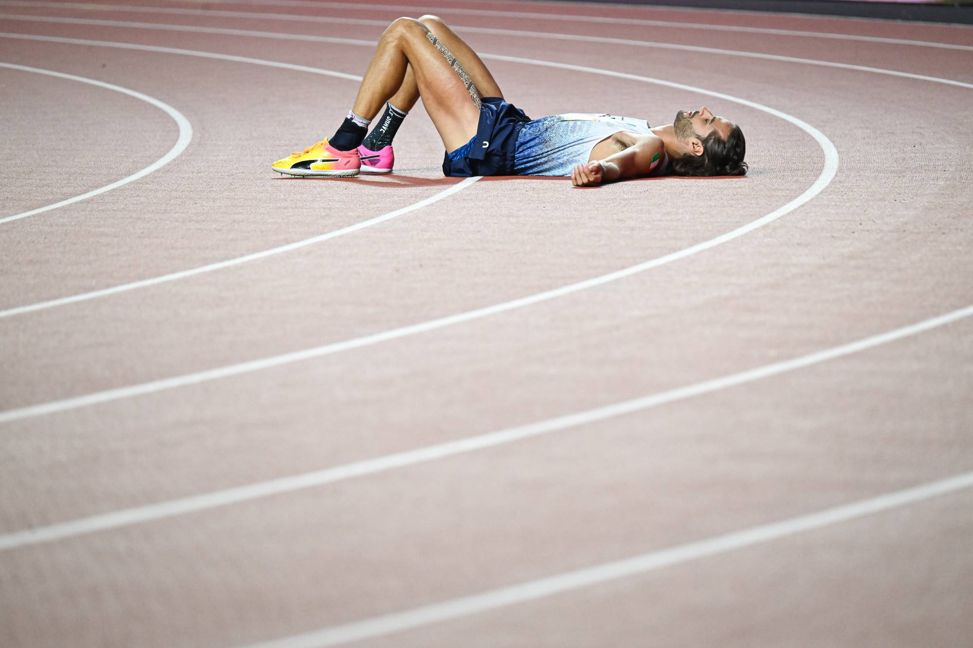 Athlete lying on a track, legs bent and arms at sides, resting after a run, wearing blue running kit and bright multicolored shoes.