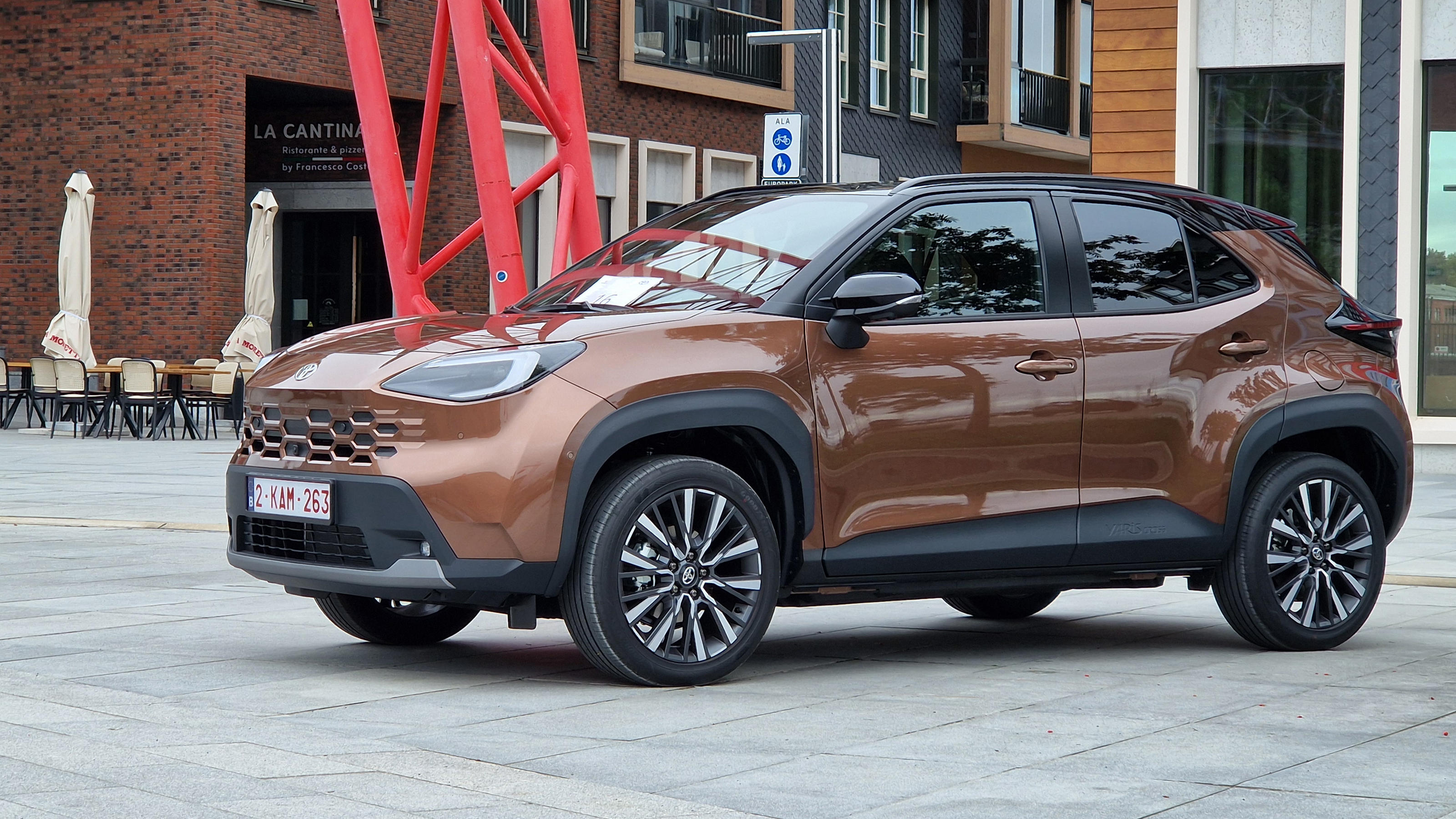 Brown SUV parked on a city plaza with red sculptural beams and outdoor seating in the background.